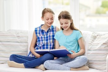 people, children, friends, literature and friendship concept - two happy girls sitting on sofa and reading book at home