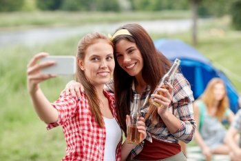 camping, travel, tourism, hike and people concept - happy young women with glass bottles drinking cider or beer and taking selfie by smartphone at camping