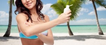 people, tanning, skincare, summer and travel concept - happy young woman in bikini swimsuit holding and applying sunscreen to her arm over tropical beach with palm trees background