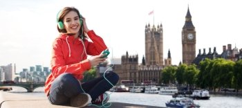 technology, travel, tourism, vacation and people concept - smiling young woman or teenage girl with smartphone and headphones listening to music over london city and thames river background