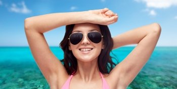 people, summer holidays, travel, tourism and beach concept - happy young woman in sunglasses and pink swimsuit over sea and blue sky background
