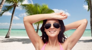 people, summer holidays, travel and tourism concept - happy young woman in sunglasses and pink swimsuit over tropical beach with palm trees background