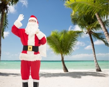 christmas, holidays, gesture and people concept - man in costume of santa claus with bag waving hand over tropical beach background