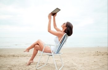 summer vacation, tourism, travel, holidays and people concept - smiling young woman with tablet pc computer sitting folding chair and taking selfie on beach