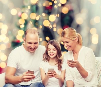 family, holidays, technology and people - smiling mother, father and little girl with smartphones over christmas tree lights background