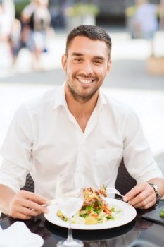 people, holidays, food and leisure concept - happy man with fork and knife eating salad for dinner at restaurant terrace