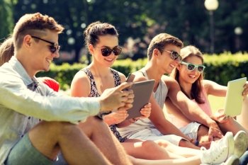 friendship, leisure, summer, technology and people concept - group of smiling friends with tablet pc computers and smartphone sitting on grass in park