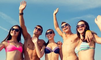 friendship, sea, holidays, gesture and people concept - group of smiling friends wearing swimwear and sunglasses waving hands on beach
