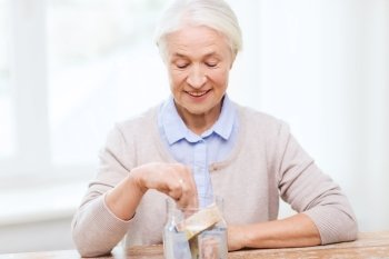 savings, money, annuity insurance, retirement and people concept - smiling senior woman putting bank notes into glass jar at home