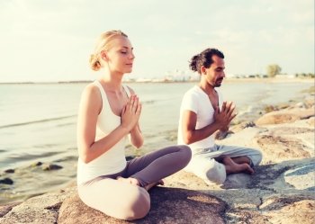fitness, sport, friendship and lifestyle concept - smiling couple making yoga exercises sitting on sand outdoors