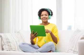 people, technology and leisure concept - happy african american young woman sitting on sofa with tablet pc computer and headphones listening to music at home