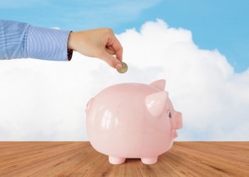 business, finance, investment, money saving and budget concept - close up of hand putting coin into piggy bank over blue sky and wooden floor background