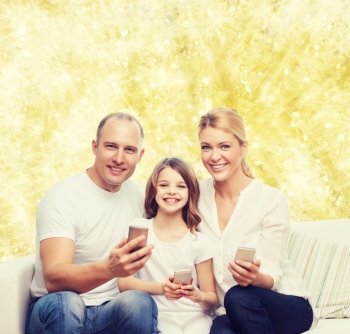 family, holidays, technology and people concept - smiling mother, father and little girl with smartphones over yellow lights background
