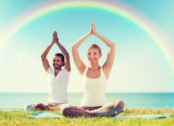 sport, fitness, yoga and people concept - smiling couple meditating and sitting on mats with raised hands over sea and rainbow in blue sky background