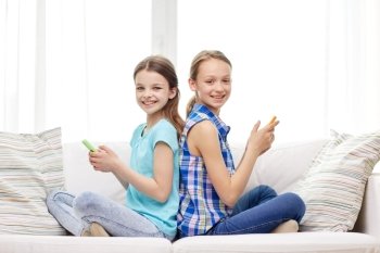 people, children, technology, friends and friendship concept - happy little girls with smartphones sitting on sofa back to back at home