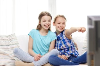 people, children, television, friends and friendship concept - two happy little girls watching tv and pointing finger at home