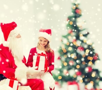 holidays, childhood and people concept - smiling little girl, santa claus with gifts over living room, christmas tree and snow background