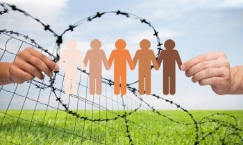 crime, imprisonment, refugee and humanity concept - multiracial couple hands holding chain of paper people pictogram over sky, grass and barb wire background