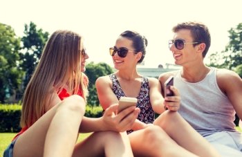 friendship, leisure, summer, technology and people concept - group of smiling friends with smartphones sitting on grass and talking in park