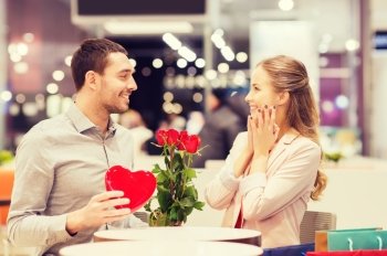 love, romance, valentines day, couple and people concept - happy young man with red flowers giving present to smiling woman at cafe in mall