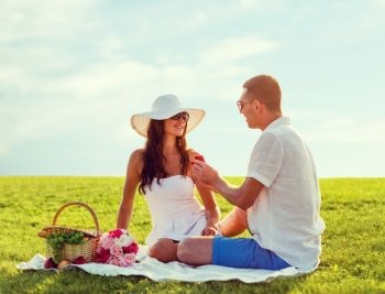 love, dating, people, proposal and holidays concept - smiling young man giving small red gift box with wedding ring to his girlfriend on picnic over blue sky and grass background
