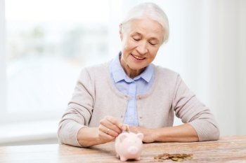 savings, money, annuity insurance, retirement and people concept - smiling senior woman putting coins into piggy bank at home