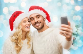 christmas, holidays, technology and people concept - happy couple in santa hats taking selfie picture with smartphone at home over blue lights background