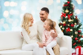 family, christmas, holidays and people concept - happy mother, father and little daughter sitting on sofa at home over blue lights background