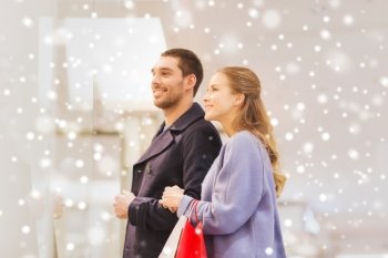 sale, consumerism and people concept - happy young couple with shopping bags looking to shopwindow in mall with snow effect