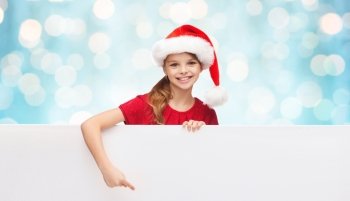 christmas, people, children and advertisement concept - happy girl child in santa helper hat pointing finger on blank white board over blue holidays lights background