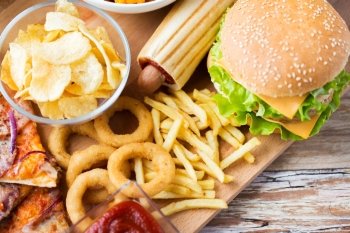 fast food and unhealthy eating concept - close up of hamburger or cheeseburger, deep-fried squid rings, french fries hotdog and potato chips on wooden table top view