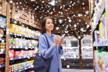 sale, shopping, consumerism and people concept - happy young woman taking notes to notebook in grocery store or market over snow effect