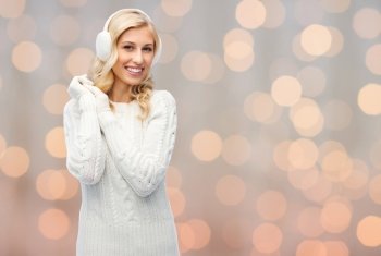 winter, fashion, christmas and people concept - smiling young woman in earmuffs and sweater over holidays lights background