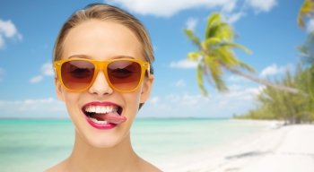 people, expression, summer vacation, travel and fashion concept - smiling young woman in sunglasses with pink lipstick on lips showing tongue over tropical beach with palm background