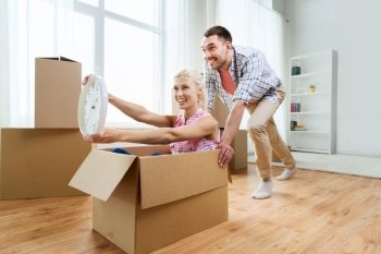 home, people, moving, time and real estate concept - happy couple having fun and riding in cardboard boxes with clock at new home