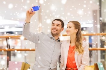sale, consumerism, technology and people concept - happy young couple with shopping bags and smartphone taking selfie in mall with snow effect