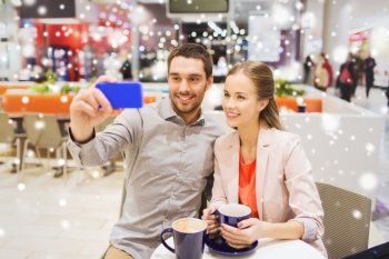 sale, shopping, consumerism, technology and people concept - happy young couple with smartphone taking selfie at cafe in mall with snow effect