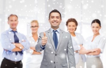 business, people, gesture and office concept - happy businessman with team showing thumbs up over office background and snow effect