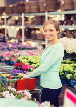 people, gardening, shopping, sale and consumerism concept - happy woman with trolley buying flowers at greenhouse or shop