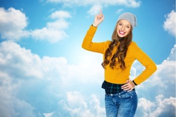 people, style and fashion concept - happy young woman or teen girl in casual clothes and hipster hat pointing finger up over blue sky and clouds background