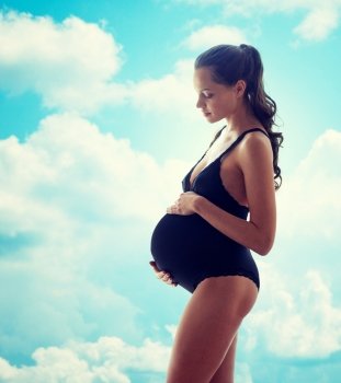 pregnancy, motherhood, people and expectation concept - happy pregnant woman in black underwear over blue sky and clouds background