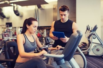 sport, fitness, lifestyle, technology and people concept - woman and trainer with tablet pc computer working out on exercise bike in gym