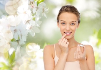 beauty, people and lip care concept - smiling young woman applying lip balm to her lips over green natural cherry blossom background