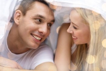 people, love, relations and bedtime concept - happy couple under blanket in bed over lights background