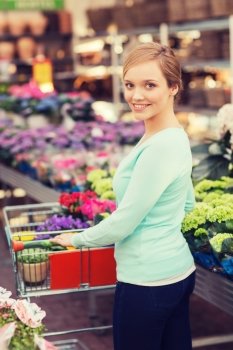 people, gardening, shopping, sale and consumerism concept - happy woman with trolley buying flowers at greenhouse or shop