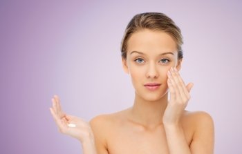beauty, people, cosmetics, skincare and health concept - young woman applying cream to her face over violet background
