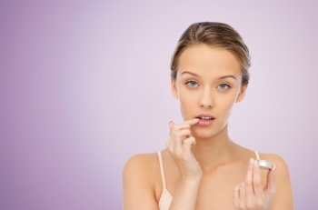 beauty, people and lip care concept - young woman applying lip balm to her lips over violet background