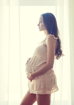 pregnancy, motherhood, people and expectation concept - happy pregnant woman in chemise near window at home