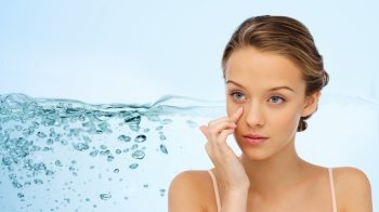 beauty, people, cosmetics, moisturizing and skin care concept - young woman applying cream to her face over water splash background