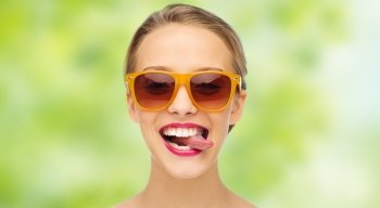 people, expression, joy and fashion concept - smiling young woman in sunglasses with pink lipstick on lips showing tongue over green summer background